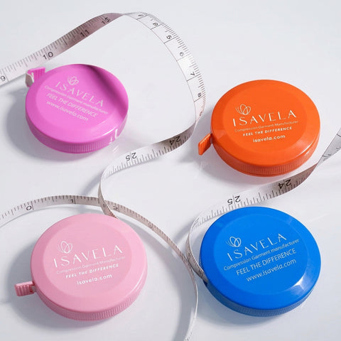 Four round plastic measuring tape cases in pink, orange, and blue, arranged on a light gray background with white measuring tapes draped around them. Each case is branded ISAVELA.